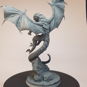 May include: A gray, detailed miniature dragon figurine with wings spread wide, perched on a rocky base. The dragon has a long, curled tail and intricate scales.