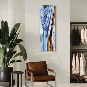 May include: Abstract painting of a wood grain pattern in shades of blue, white, and orange. The painting is framed and hanging on a wall in a room with a brown leather armchair and a small table.