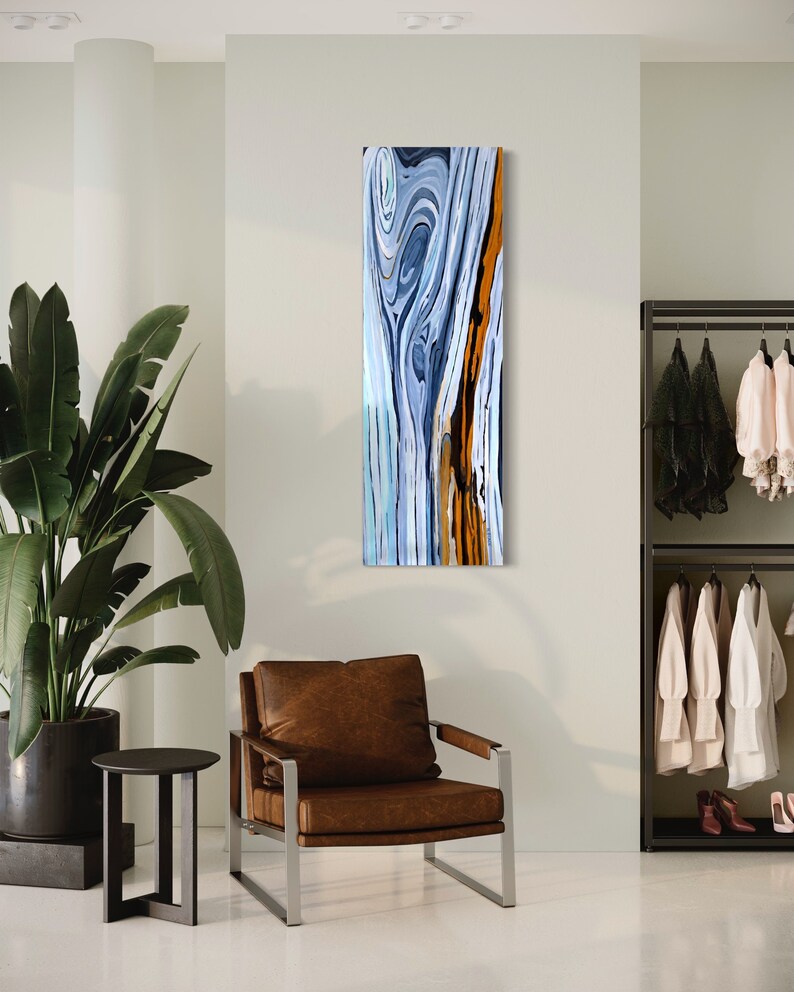 May include: Abstract painting of a wood grain pattern in shades of blue, white, and orange. The painting is framed and hanging on a wall in a room with a brown leather armchair and a small table.