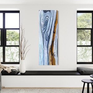 May include: Abstract painting with swirling blue, white, and orange colors. The painting has a vertical format and is framed in a modern, minimalist style.