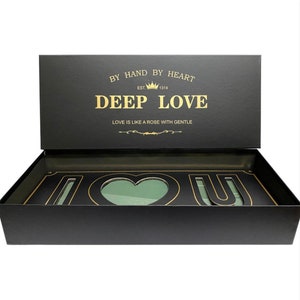 May include: Black gift box with a gold inscription that reads "By Hand By Heart Est. 1314 Deep Love Love is like a rose with gentle". The box is open and reveals a black and gold "I Love You" sign with a heart-shaped cutout in the middle.