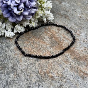 May include: A delicate black beaded necklace. The necklace is composed of small, round, black beads. The necklace is displayed on a textured, gray surface, with a bouquet of purple and white flowers in the background.