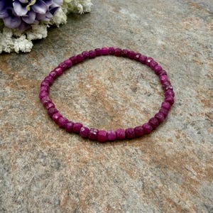 May include: A vibrant, beaded bracelet composed of small, faceted, deep pink gemstones. The bracelet is circular and rests on a textured, neutral-toned surface. The gemstones are evenly sized and closely spaced, creating a continuous band of color.