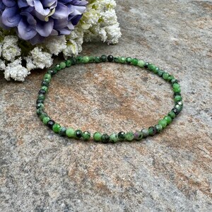 May include: A beaded bracelet featuring small, round, green and black beads. The bracelet is made of natural stones and is displayed on a textured, gray surface. The beads have a faceted cut, adding sparkle.