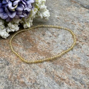 May include: A delicate necklace composed of small, round, yellow beads. The necklace is displayed on a textured, multi-toned stone surface, with a bouquet of purple and white flowers in the background. The beads appear to be approximately 0.12 inches in diameter.