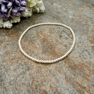 May include: A delicate, white pearl necklace. The necklace is a single strand of small, round pearls. The pearls are strung together to form a complete circle. The necklace is displayed on a textured, stone-like surface.