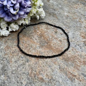May include: A delicate black beaded necklace. The necklace is composed of small, faceted black beads strung together. The beads are arranged in a single strand, forming a circular shape. The necklace is displayed on a textured, gray surface, with purple and white flowers in the background.