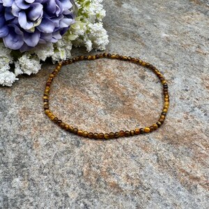 May include: A delicate bracelet made of small, round tiger's eye beads. The bracelet is a warm brown color with golden and black accents. The beads are strung together to form a circular shape. The bracelet is displayed on a textured, gray surface.