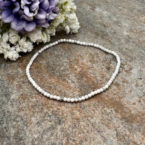 May include: A delicate white beaded necklace with subtle gray accents. The necklace is composed of small, round beads, creating a simple yet elegant design. The necklace is displayed on a textured, neutral-toned surface, with a floral arrangement in the background.