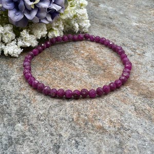 May include: A vibrant, faceted ruby bracelet. The bracelet is composed of small, round, deep pink gemstones strung together. The bracelet is displayed on a textured, gray surface, with a bouquet of white and purple flowers in the background.