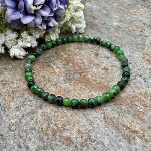 May include: A beaded bracelet featuring small, round, green and black beads. The bracelet is strung together, forming a circular shape. The beads have a polished appearance. The background includes a stone surface and a cluster of purple and white flowers.