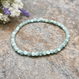 May include: A delicate bracelet composed of small, round, pale green beads. The beads are strung together to form a flexible band. The bracelet is displayed on a textured, neutral-toned surface, with a hint of floral accents in the background.