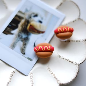 Puede incluir: Dos pequeños pendientes de arcilla polimérica en forma de perrito caliente de color marrón y rojo con detalle de mostaza amarilla.
