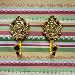 Puede incluir: Dos ganchos decorativos de tono dorado con diseños florales ornamentados. Los ganchos están montados sobre un fondo a rayas.