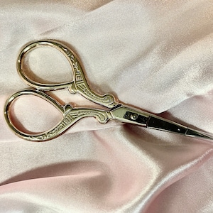 May include: Gold-toned decorative scissors with intricate details on the handles. The scissors have a sharp, pointed blade and a small, clear plastic tip.
