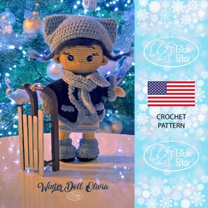 Puede incluir: Patrón de crochet para una muñeca de invierno llamada Olivia. La muñeca lleva un gorro azul, una bufanda azul y un suéter a rayas azul y blanco. La muñeca sostiene un trineo de madera. El patrón está disponible para comprar en Etsy.