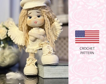 Könnte beinhalten: Eine gehäkelte Engelpuppe mit blonden Haaren, die einen weißen Barett, ein weißes Kleid und weiße Schuhe trägt. Die Puppe steht auf einer Holzkiste mit den Worten "Angel Michalina" darunter. Der Hintergrund ist ein rosa Muster mit weißen Federn.