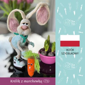 May include: A crocheted bunny rabbit holding a carrot. The bunny is wearing a light blue shirt and white sneakers. The carrot has a face and green hair. The background is a light blue with a white pattern of carrots. The text "WZÓR SZYDŁKOWY" is in the background.