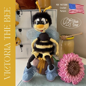 Könnte beinhalten: Handgefertigte Häkelpuppe Biene namens "Victoria the Bee" mit schwarzen und gelben Streifen, blauen Schuhen und einer rosa Blume. Die Puppe sitzt auf einem Honigtopf. Das Bild enthält auch den Text "PDF Pattern Crochet Pattern" und die amerikanische Flagge.