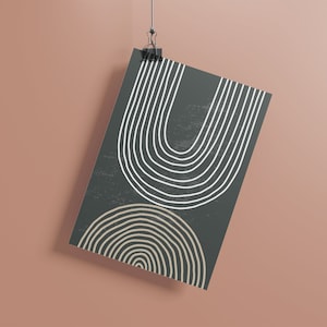 Puede incluir: Una impresión artística rectangular con un diseño minimalista. La impresión tiene un fondo gris oscuro con líneas curvas blancas y beige dispuestas en patrones semicirculares. La impresión está sujeta por un clip negro sobre un fondo melocotón.