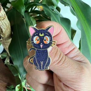 May include: A gold-colored pin featuring a cartoon cat with a crescent moon on its forehead. The cat has large orange eyes, pink ears, and a dark blue body with glitter. The pin is held in front of a green leafy background.