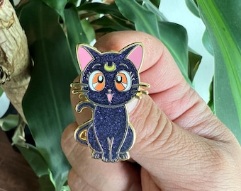 Peut inclure: Épingle dorée représentant un chat de dessin animé avec un croissant de lune sur le front. Le chat a de grands yeux orange, des oreilles roses et un corps bleu foncé pailleté. L'épingle est présentée devant un fond de feuillage vert.
