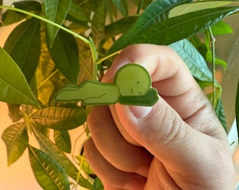 May include: A green enamel pin featuring a cartoon character lying down. The pin has a rounded head and a body resting on a cloud-like shape. The pin is held in front of a background of green leaves.