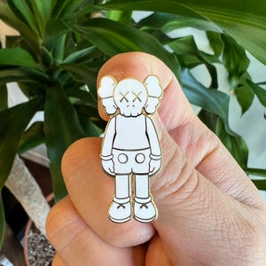 May include: White enamel pin featuring a cartoon character with a simple design. The character has a round head with two small bumps on top, X-shaped eyes, and a small mouth. The character is wearing shorts and has short legs and arms.