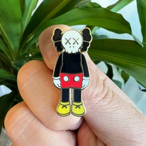 May include: A black and white enamel pin featuring a cartoon character with a skull face and red shorts. The character has black hair and yellow shoes. The pin is gold-toned.