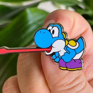 Puede incluir: Un pin de esmalte azul y blanco de un dinosaurio de dibujos animados con una larga lengua roja que sobresale. El dinosaurio lleva un atuendo morado y amarillo.