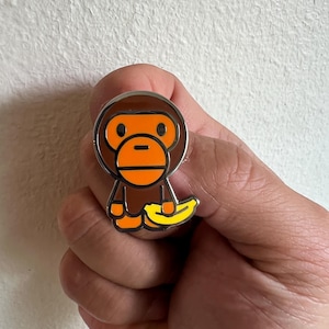 Pode incluir: Um alfinete de macaco castanho e laranja com uma banana amarela. O alfinete tem uma borda prateada. O macaco tem uma cabeça redonda e uma boca retangular. A banana é curvada.