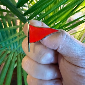以下が含まれることがあります： 金色の縁とポストが付いた赤いエナメル旗ピン。 ピンは三角形で、緑の葉を背景に手で持っています。