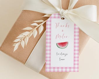 Peut inclure: Un cadeau emballé avec un ruban crème et une étiquette vichy rose. L'étiquette indique "Thanks a Melon" avec une illustration de tranche de pastèque. Une brindille de fleurs séchées est également présente.