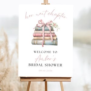 May include: A white sign on an easel with the text "her next chapter" in a delicate script. Below, a stack of books is adorned with a pink ribbon and flowers. The sign reads "Welcome to Amber's Bridal Shower" with the date 08.10.2026.