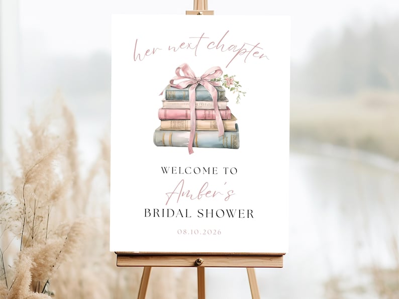 May include: A white sign on an easel with the text "her next chapter" in a delicate script. Below, a stack of books is adorned with a pink ribbon and flowers. The sign reads "Welcome to Amber's Bridal Shower" with the date 08.10.2026.