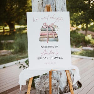 May include: A white sign on a wooden easel with the text "her next chapter" and "Welcome to Amber's Bridal Shower". The sign features a watercolour illustration of a stack of books tied with a pink ribbon and flowers. The date 08.10.2026 is also displayed.