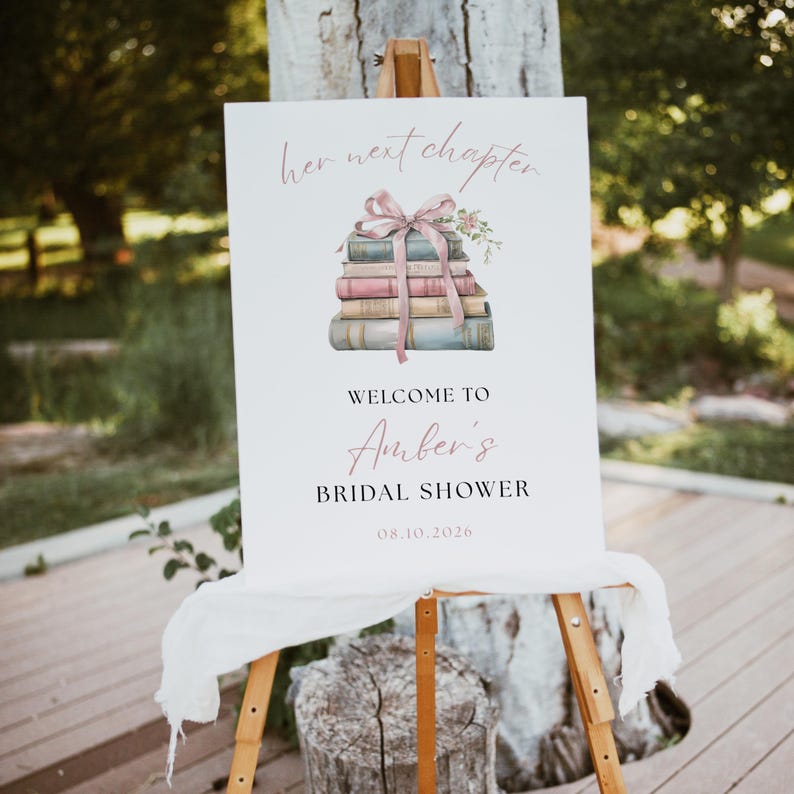 May include: A white sign on a wooden easel with the text "her next chapter" and "Welcome to Amber's Bridal Shower". The sign features a watercolour illustration of a stack of books tied with a pink ribbon and flowers. The date 08.10.2026 is also displayed.