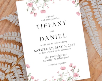 May include: A wedding invitation with the names TIFFANY and DANIEL, and the date Saturday, May 5, 2027. The invitation is white with a floral border of pink flowers and green leaves. The invitation is on a wooden tray.