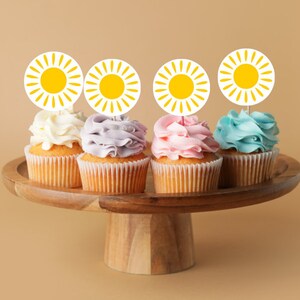 May include: Four cupcakes decorated with different colored frosting and topped with yellow sun-shaped cupcake toppers on a wooden cake stand.