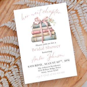 May include: A white bridal shower invitation with a pink and blue illustration of a stack of books tied with a pink ribbon. The text reads "her next chapter" and "Please join us for a Bridal Shower honouring Amber Johnson" with the date and time of the event.