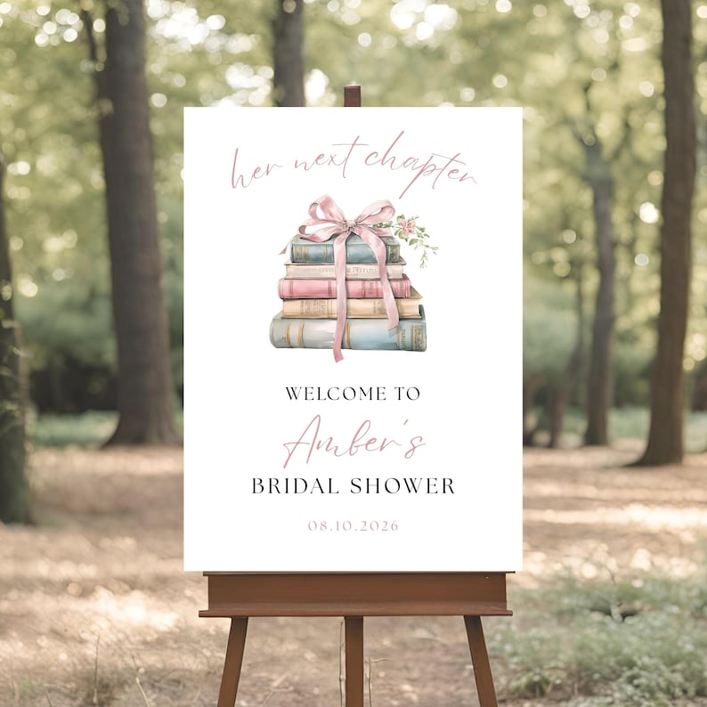 May include: A white sign on a wooden easel with the text "her next chapter" in a cursive font. Below is a stack of books tied with a pink ribbon and flowers. The sign reads "Welcome to Amber's Bridal Shower 08.10.2026".