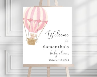 Puede incluir: Un cartel de bienvenida blanco con un globo aerostático rosa y blanco con un osito de peluche dentro. El cartel dice "Bienvenido al baby shower de Samantha el 12 de octubre de 2024".