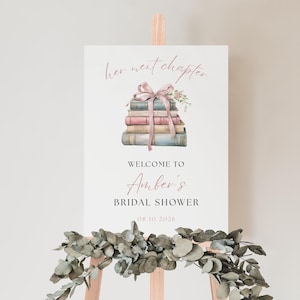 Peut inclure: Panneau blanc sur chevalet en bois avec le texte "her next chapter" et "Welcome to Amber's Bridal Shower". Le panneau présente une pile de livres liés par un ruban rose et des accents floraux. Guirlande d'eucalyptus à la base.