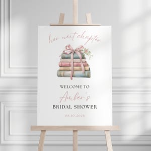 May include: A white sign on a wooden easel with the text "her next chapter" and "Welcome to Amber's Bridal Shower 08.10.2026". The sign features a watercolour illustration of a stack of books tied with a pink ribbon and floral accents.