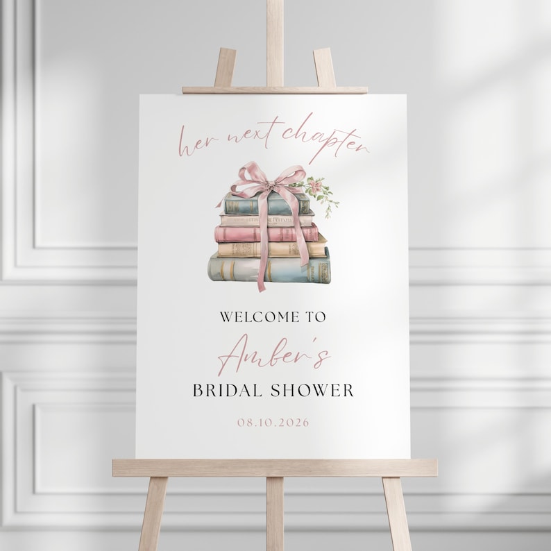 May include: A white sign on a wooden easel with the text "her next chapter" and "Welcome to Amber's Bridal Shower 08.10.2026". The sign features a watercolour illustration of a stack of books tied with a pink ribbon and floral accents.