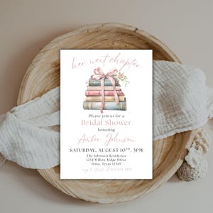 May include: A bridal shower invitation with a stack of books tied with a pink ribbon and flowers. The text reads "her next chapter" and "Please join us for a Bridal Shower honoring Amber Johnson. SATURDAY, AUGUST 10TH, 3PM. The Johnson Residence 1234 Willow Ridge Drive Irma, Texas 12345."