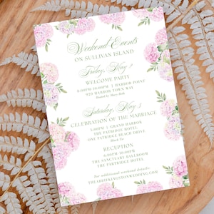 May include: A wedding invitation with the text "Weekend Events on Sullivan Island" and event details. The invitation is decorated with pink hydrangea flowers and green leaves. It rests on a wooden tray with fern accents.