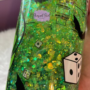 Puede incluir: Un vaso con purpurina verde con siluetas de murci&eacute;lagos negros y gr&aacute;ficos de dados. El vaso tiene el texto "Pleasant Point Designs" y opciones de personalizaci&oacute;n de bebidas. El interior est&aacute; lleno de purpurina verde y amarilla.