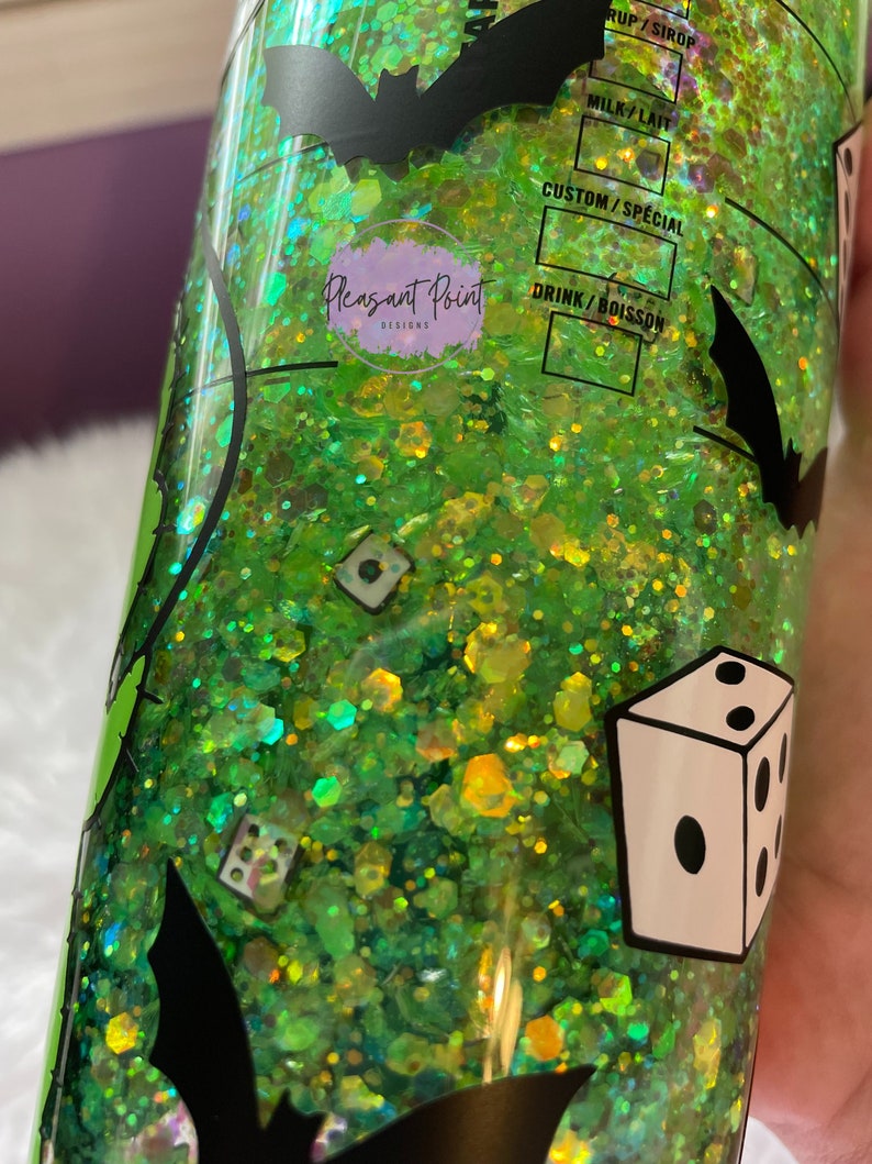 Puede incluir: Un vaso con purpurina verde con siluetas de murci&eacute;lagos negros y gr&aacute;ficos de dados. El vaso tiene el texto "Pleasant Point Designs" y opciones de personalizaci&oacute;n de bebidas. El interior est&aacute; lleno de purpurina verde y amarilla.