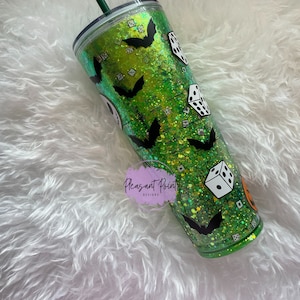 Puede incluir: Un vaso verde con purpurina y calcoman&iacute;as de murci&eacute;lagos y dados negros. El vaso tiene una pajita verde y una tapa negra. El vaso est&aacute; decorado con un tema de Halloween. El vaso es de pl&aacute;stico.